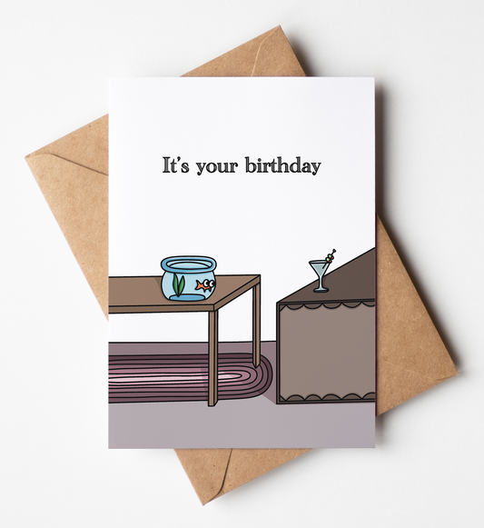 A birthday greeting card with a brown kraft envelope, featuring a minimalist illustration of a fishbowl on a table and a cocktail glass on a counter, with the text 'It's your birthday' on a white card.