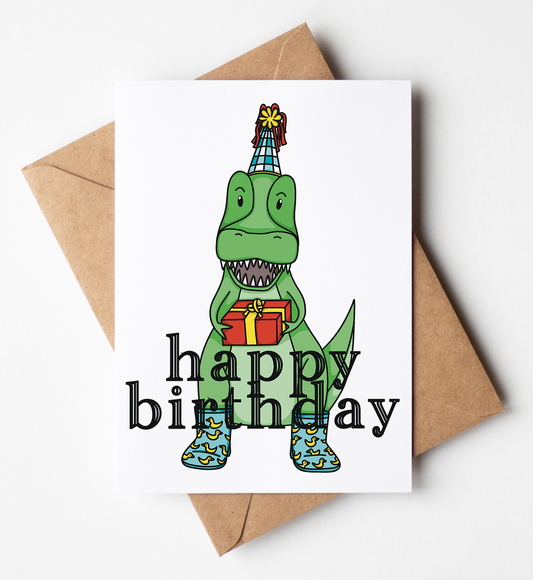 A white birthday card featuring a cartoon green dinosaur wearing a party hat and holding a gift, with the words 'happy birthday' in black. The card is shown with a brown kraft envelope on a light gray background.