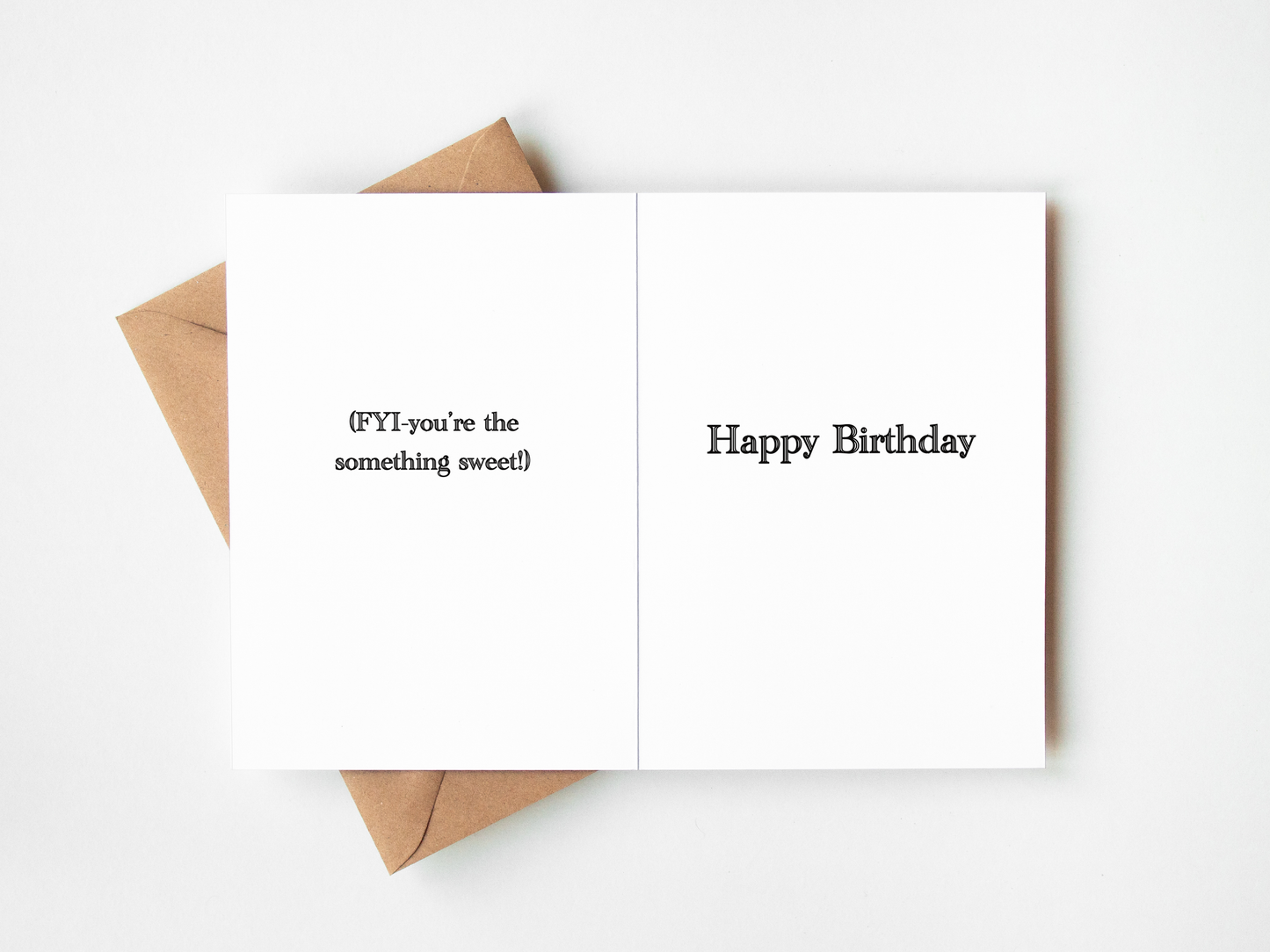 A folded white greeting card with black text reading '{FYI} you're the something sweet!' on the left panel and 'Happy Birthday' on the right panel, shown open with a brown kraft envelope behind it on a white background.