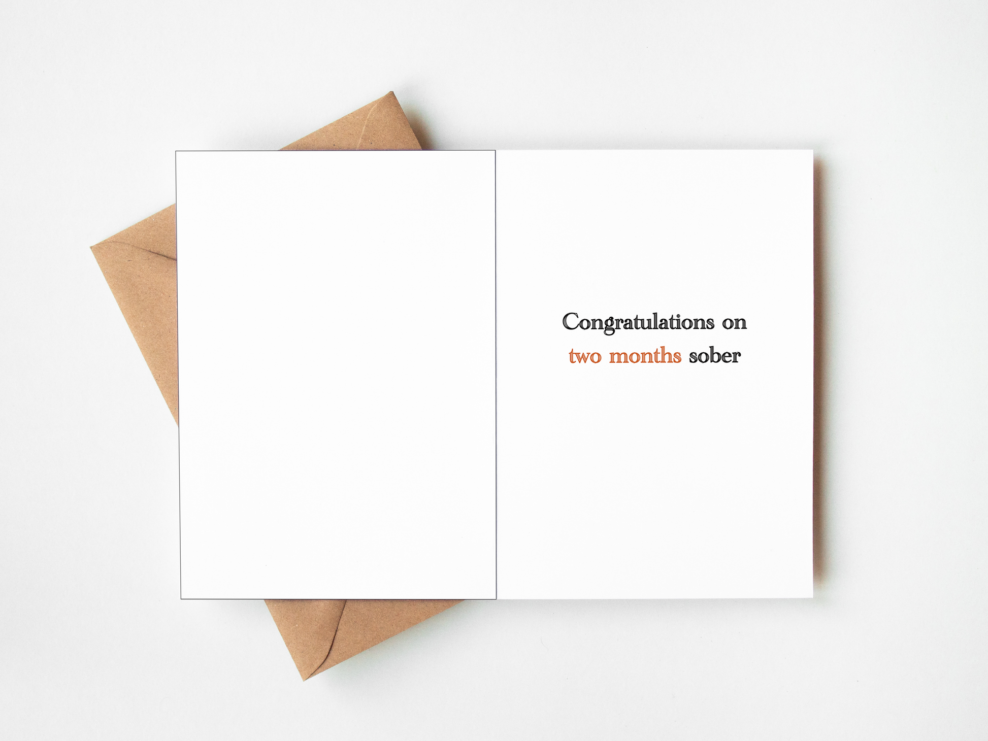 An open white greeting card with the text 'Congratulations on two months sober' printed on the right side, with a brown kraft envelope partially visible underneath on a white background.