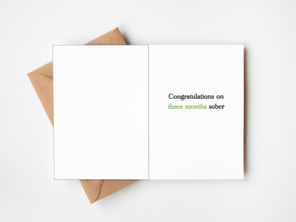 An open white greeting card with the text 'Congratulations on two months sober' printed on the right side, with a brown kraft envelope partially visible underneath on a white background.