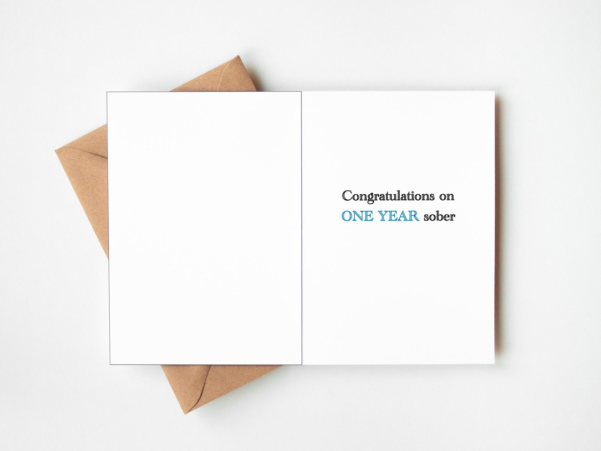 An open white greeting card with the text 'Congratulations on ONE YEAR sober' in black and blue lettering, placed on top of a brown kraft envelope against a clean white background.
