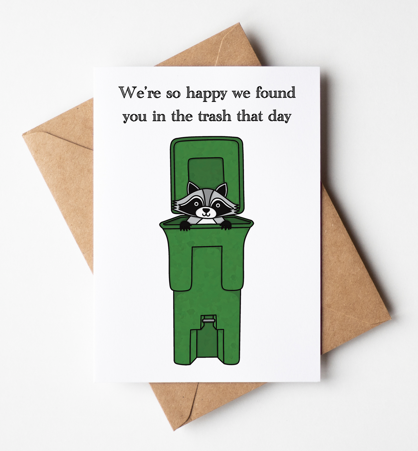 A white greeting card with a humorous cartoon of a raccoon peeking out from a green trash bin, with the text 'We're so happy we found you in the trash that day'. The card is displayed on top of a brown kraft paper envelope against a clean white background.