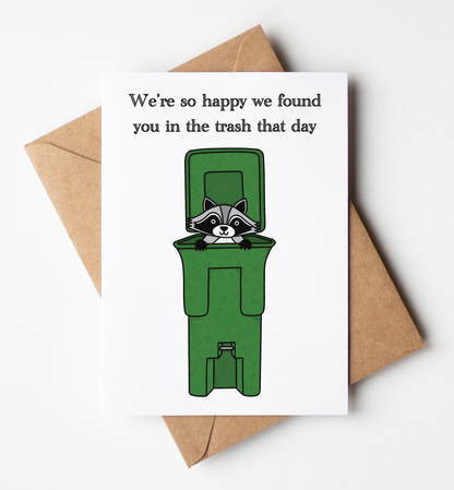 A white greeting card with a humorous cartoon of a raccoon peeking out from a green trash bin, with the text 'We're so happy we found you in the trash that day'. The card is displayed on top of a brown kraft paper envelope against a clean white background.