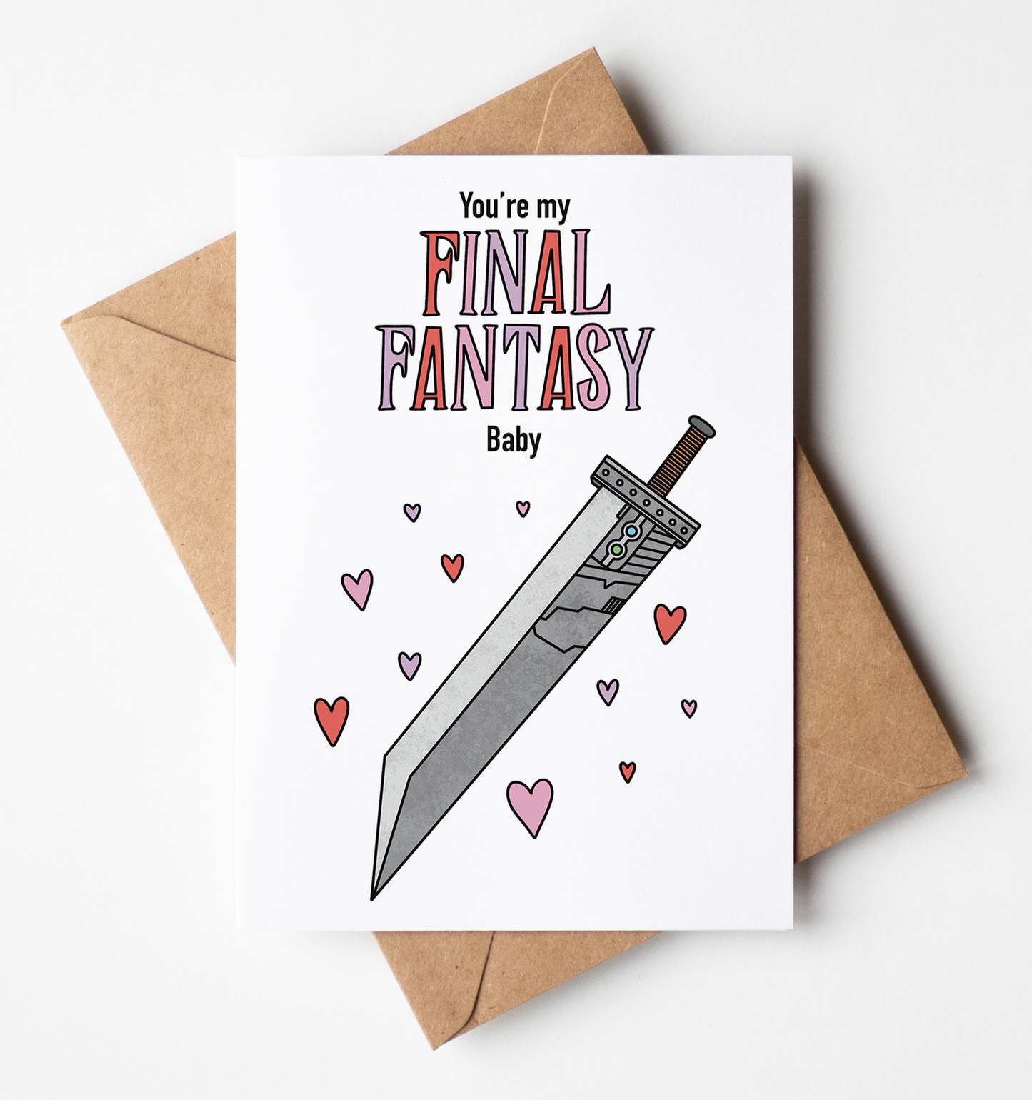 A white greeting card with a sword and heart illustration and the text 'You're my Final Fantasy Baby', placed on top of a brown kraft envelope against a light gray background.