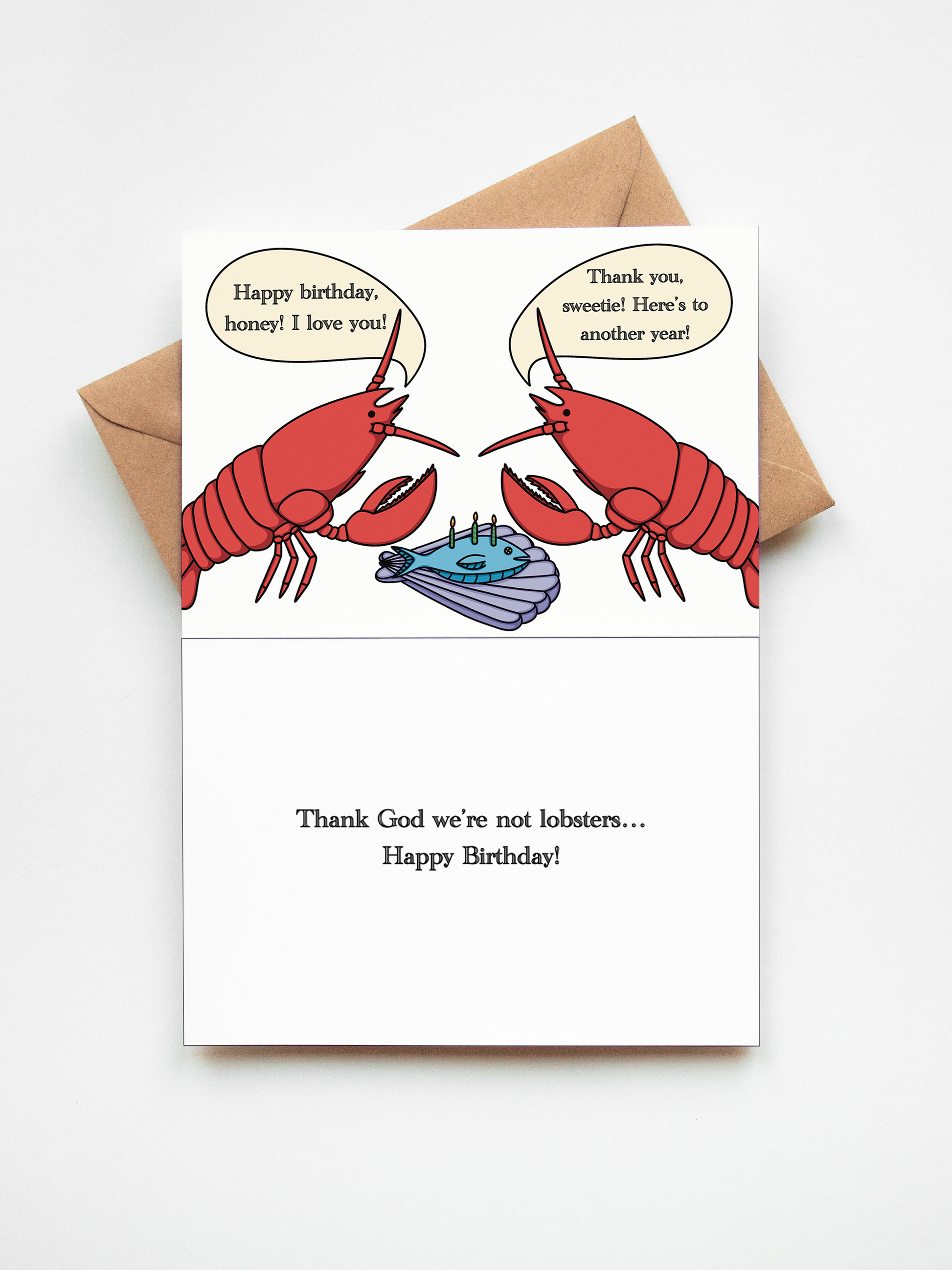 A humorous lobster birthday card shown open with a kraft envelope behind it on a white background. The left panel features a cartoon lobster and a fly with speech bubbles, and the right panel reads, 'Thank God we're not lobsters.