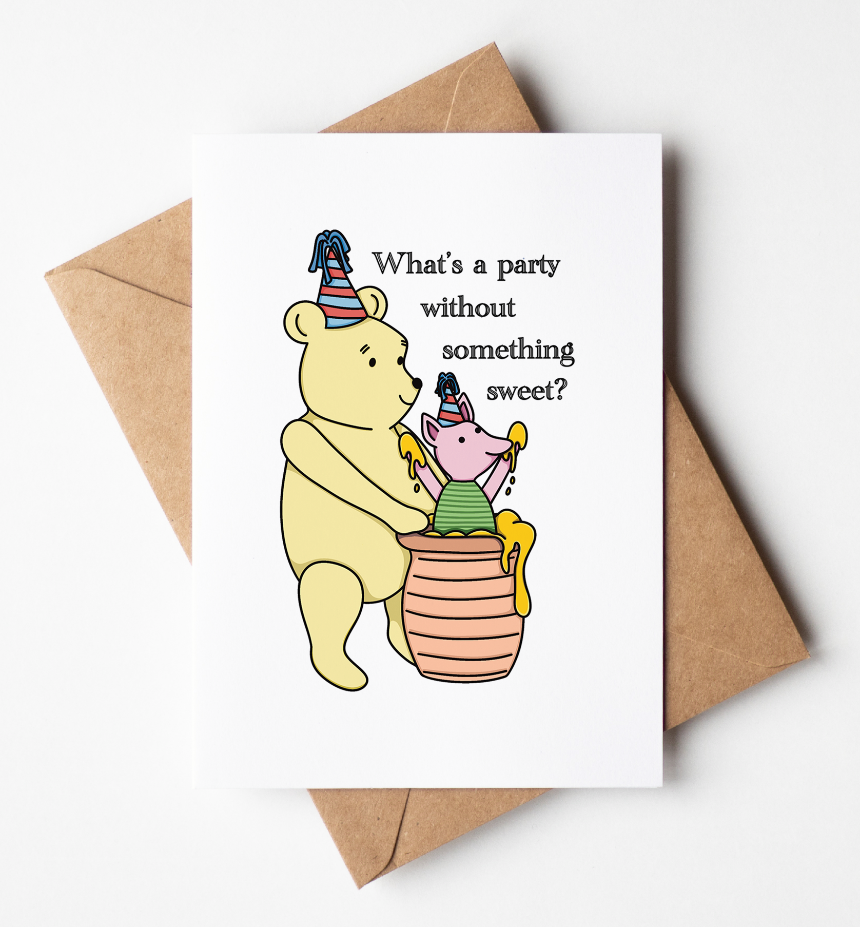 A white greeting card with a cartoon bear and pig party illustration and the text 'What's a party without something sweet?' placed on top of a brown kraft envelope against a light gray background.