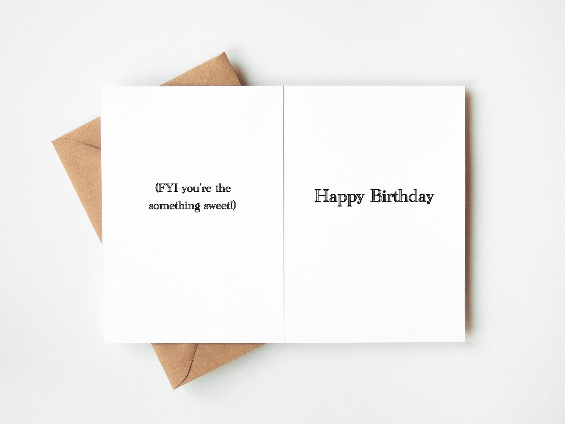 A folded white greeting card with black text reading '{FYI} you're the something sweet!' on the left panel and 'Happy Birthday' on the right panel, shown open with a brown kraft envelope behind it on a white background.