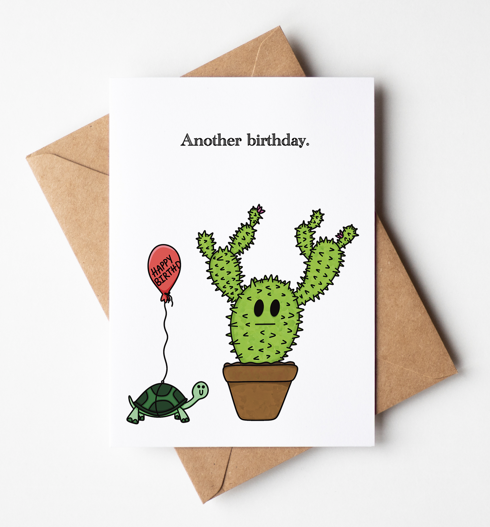 A white greeting card with the text 'Another birthday.' and a cute illustration of a green cactus in a pot holding a red balloon, placed on top of a brown kraft envelope against a light gray background.