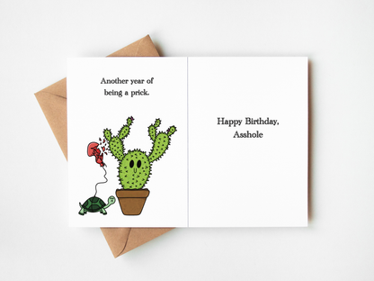 A novelty birthday card shown open on a white background with a brown envelope behind it. The left panel features a cartoon cactus in a pot with a red balloon and a turtle, with the text 'Another year of being a prick.
