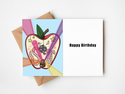 An open greeting card with a white right panel reading 'Happy Birthday' and a colorful left panel showing a stylized apple filled with party-themed illustrations, styled with brown envelopes on a light gray background.